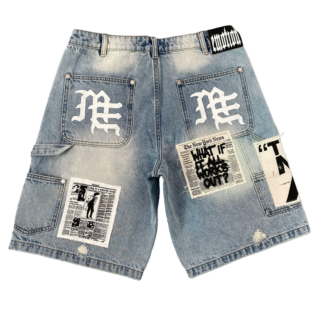 Light blue denim shorts with a heavily distressed wash, featuring white gothic lettering on the back pockets, a newspaper patch on the right leg, and a graphic patch on the left leg, shown against a plain white background.